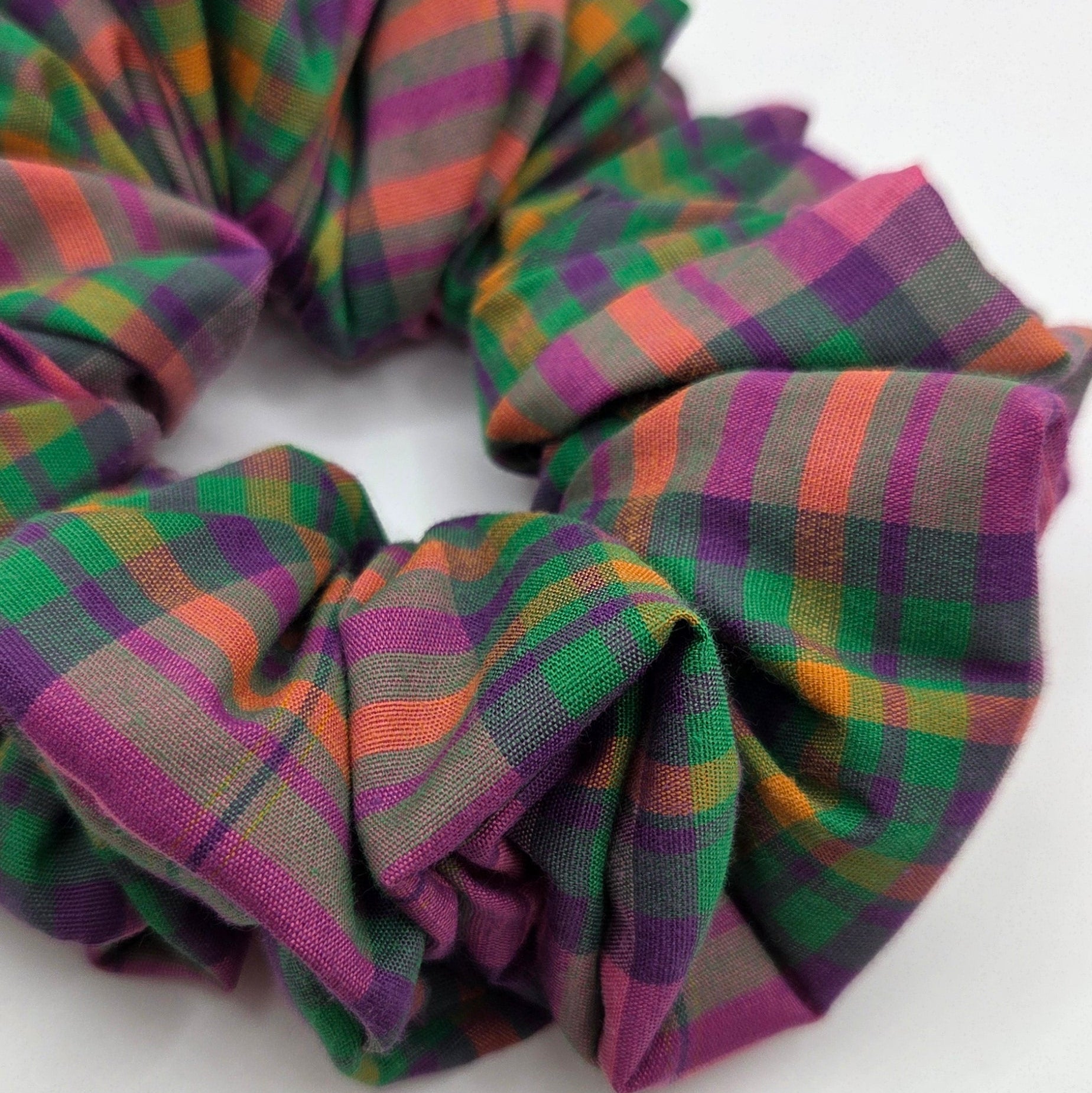 A fabric scrunchie with a multicolor plaid pattern featuring green, purple, orange, and pink, shown on a plain white background.