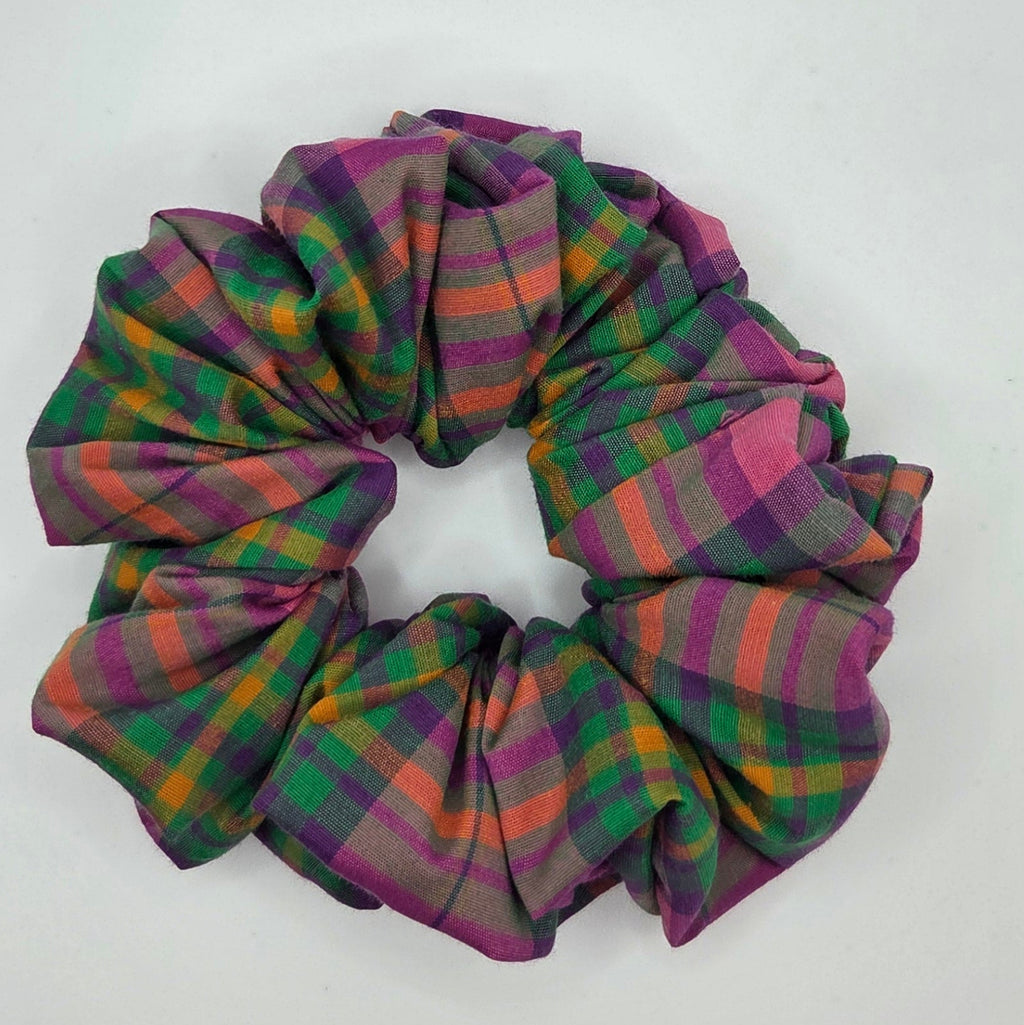 A fabric hair scrunchie with a multicolor plaid pattern in purple, green, orange, and pink, resting on a plain white surface.