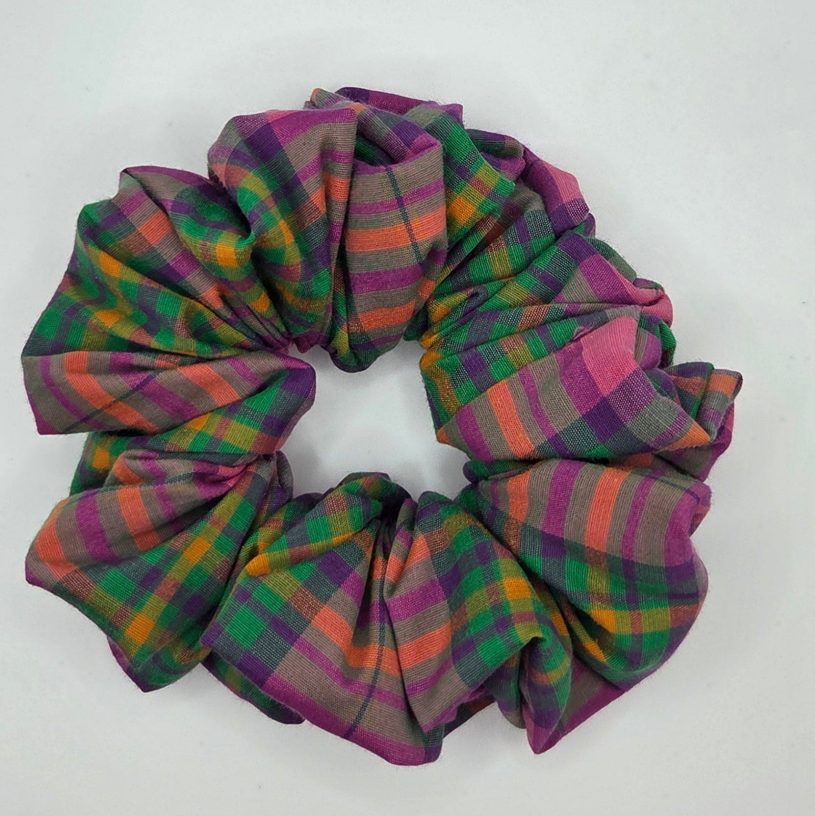 A fabric hair scrunchie with a multicolor plaid pattern in purple, green, orange, and pink, resting on a plain white surface.
