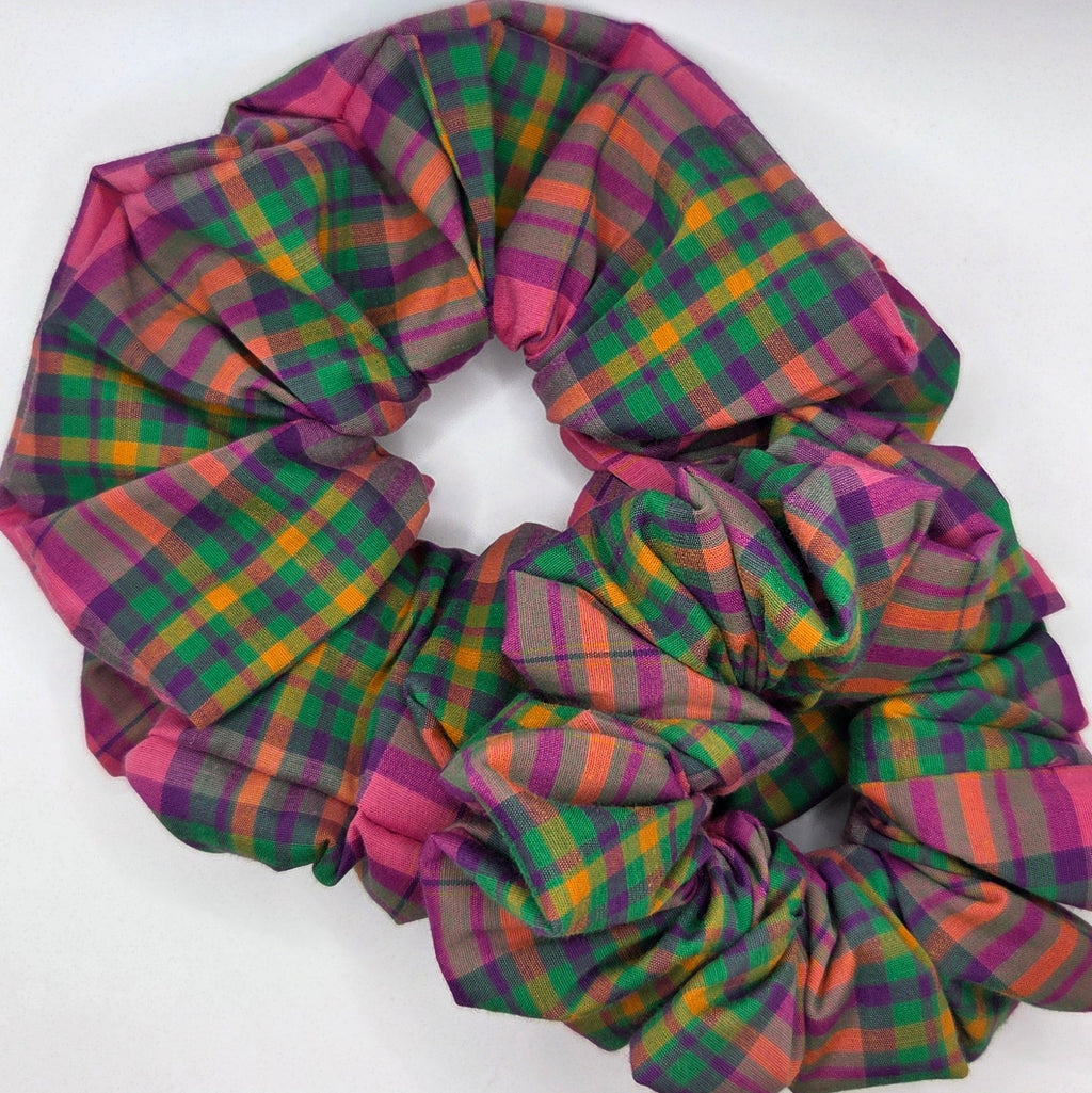 A fabric hair scrunchie with a multicolor tartan plaid pattern in green, pink, purple, and yellow, displayed on a white surface.