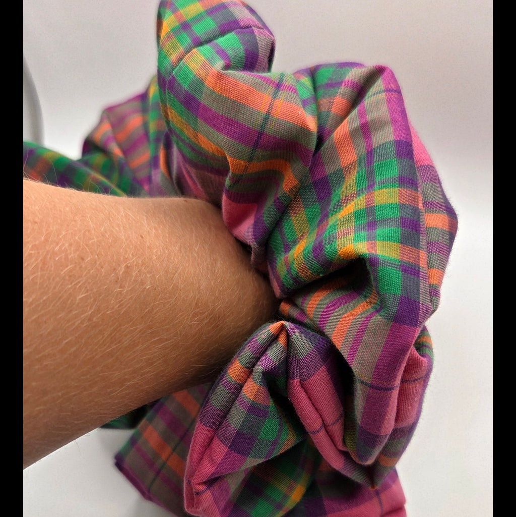 A fabric hair scrunchie with a multicolor plaid pattern in pink, green, orange, and purple, shown being worn on a person's wrist against a plain white background.