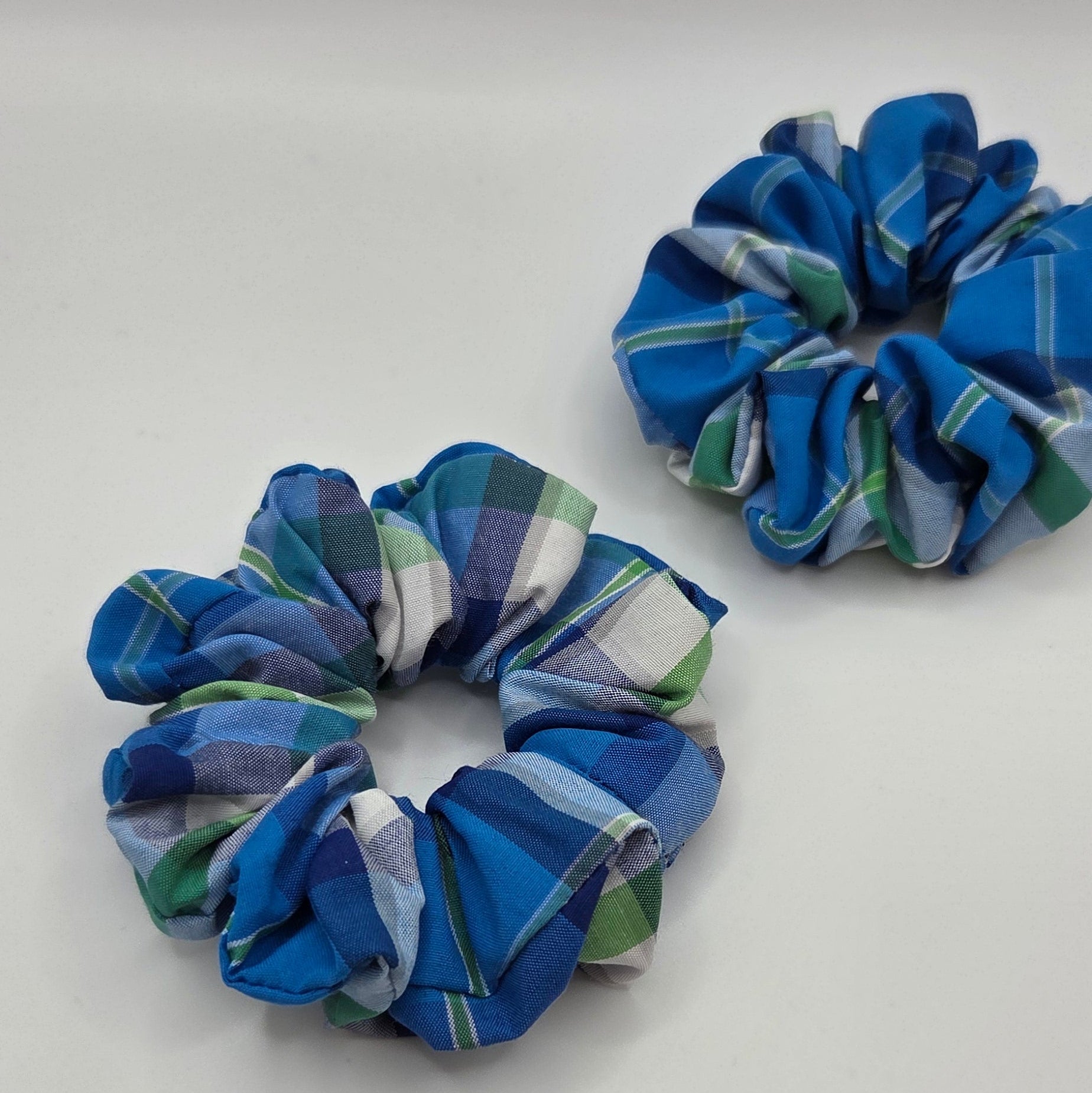 Two fabric hair scrunchies with a blue, green, and white plaid pattern, displayed on a plain light gray background.