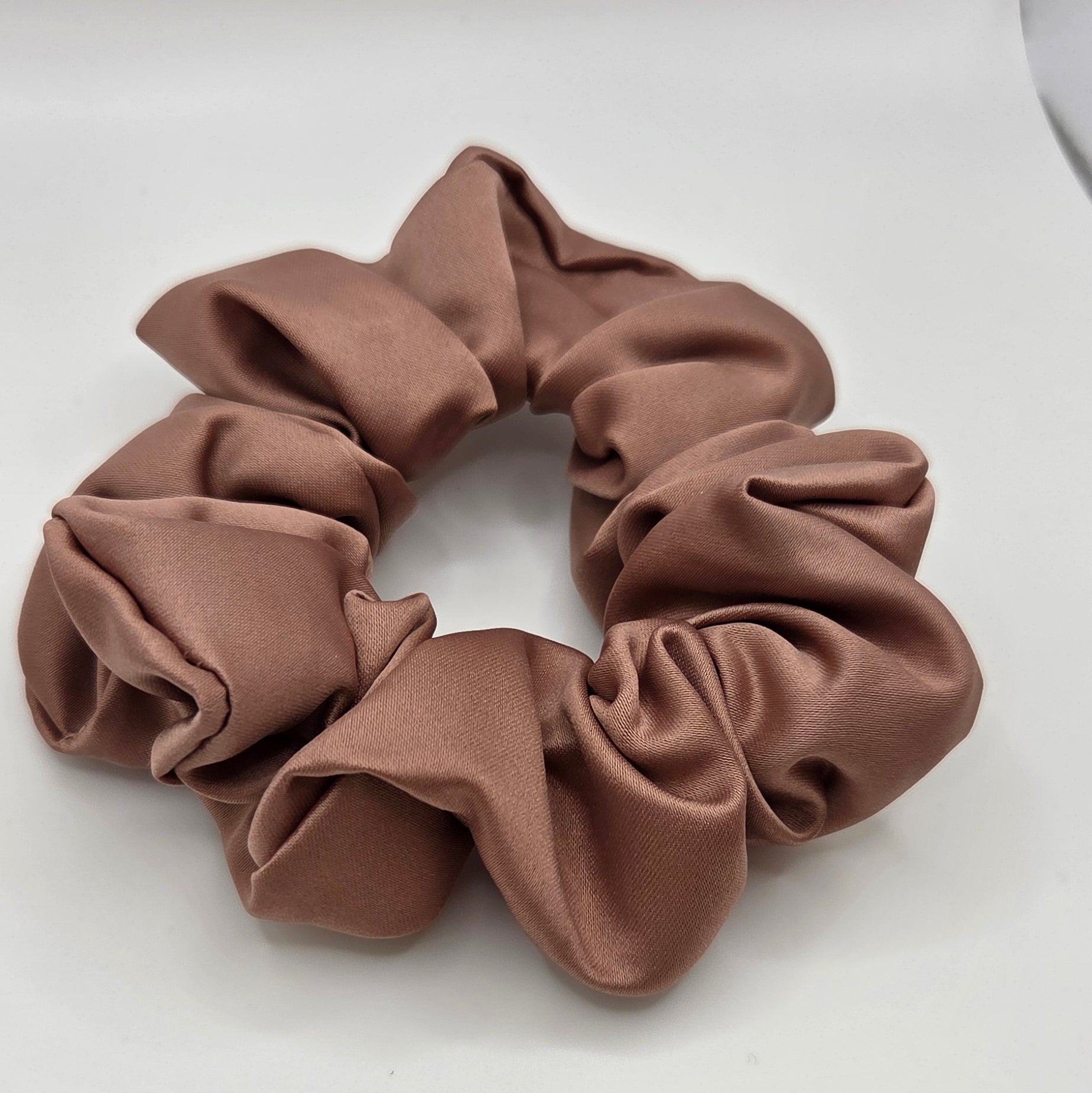 A single brown fabric scrunchie with a soft, slightly shiny satin-like finish, photographed on a plain light gray background.