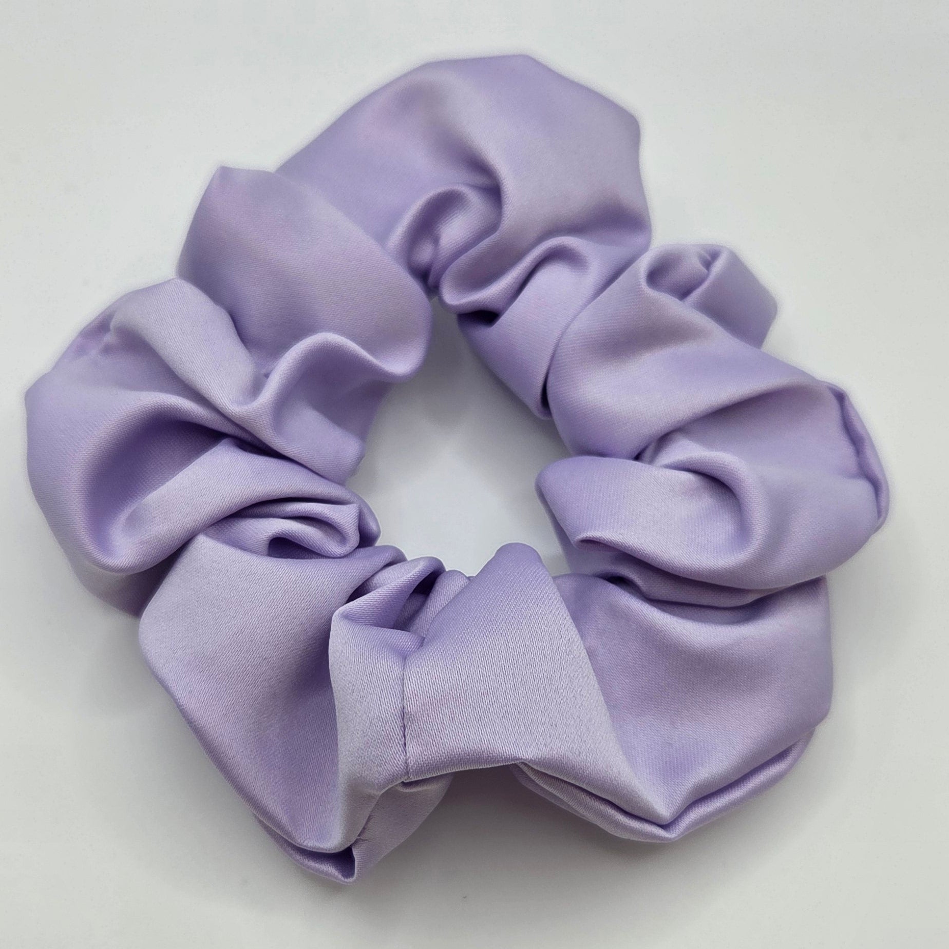 A lavender purple fabric scrunchie with a soft satin-like sheen, photographed on a plain light gray background.