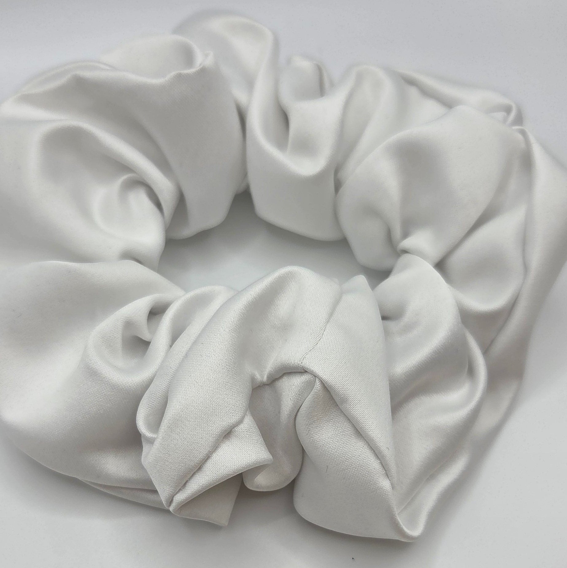 A white satin or silk scrunchie with a soft, draped texture, photographed on a plain light gray background.
