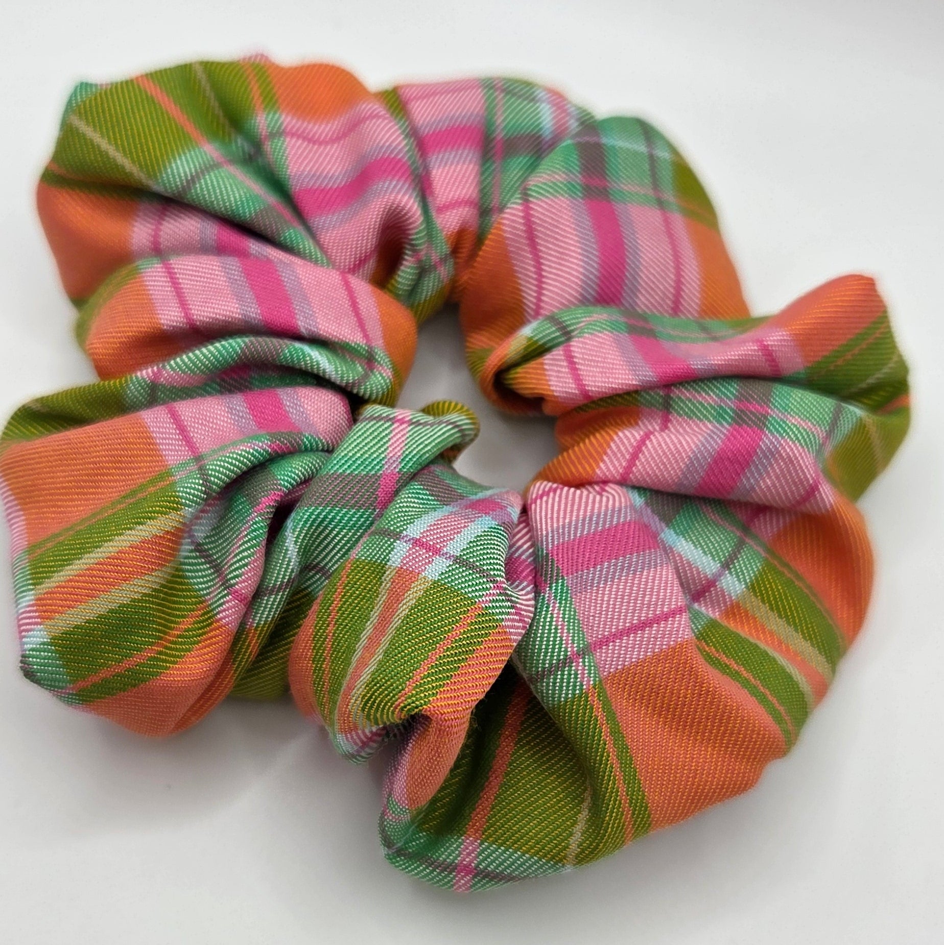 A fabric scrunchie with a plaid pattern in orange, green, pink, and white, photographed on a plain light gray background.