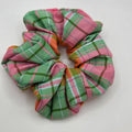 A fabric scrunchie with a pink, green, and white plaid pattern, photographed on a plain light gray background.