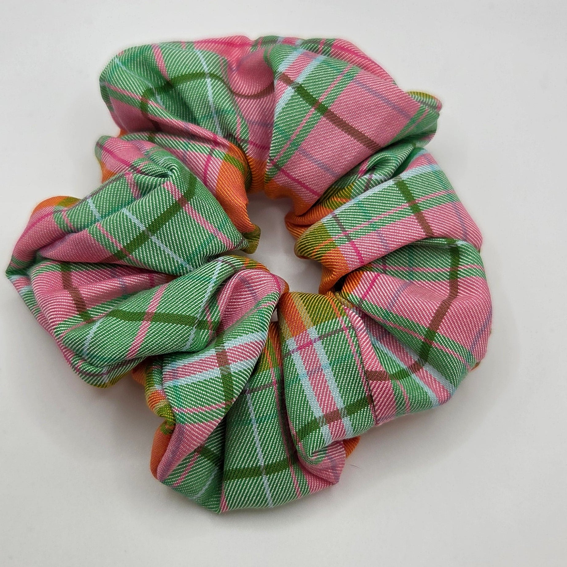 A fabric scrunchie with a pink, green, and white plaid pattern, photographed on a plain light gray background.