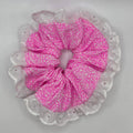 A pink fabric scrunchie with a small white floral pattern, layered over a white lace trim, shown against a plain light gray background.