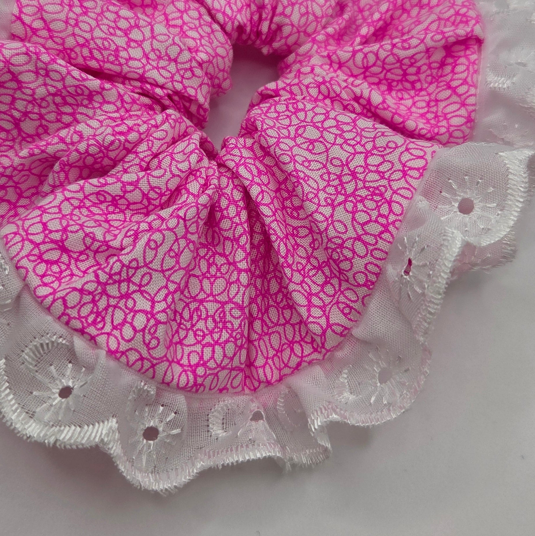 A pink fabric scrunchie with a white floral pattern and white lace trim, shown on a plain light gray background.