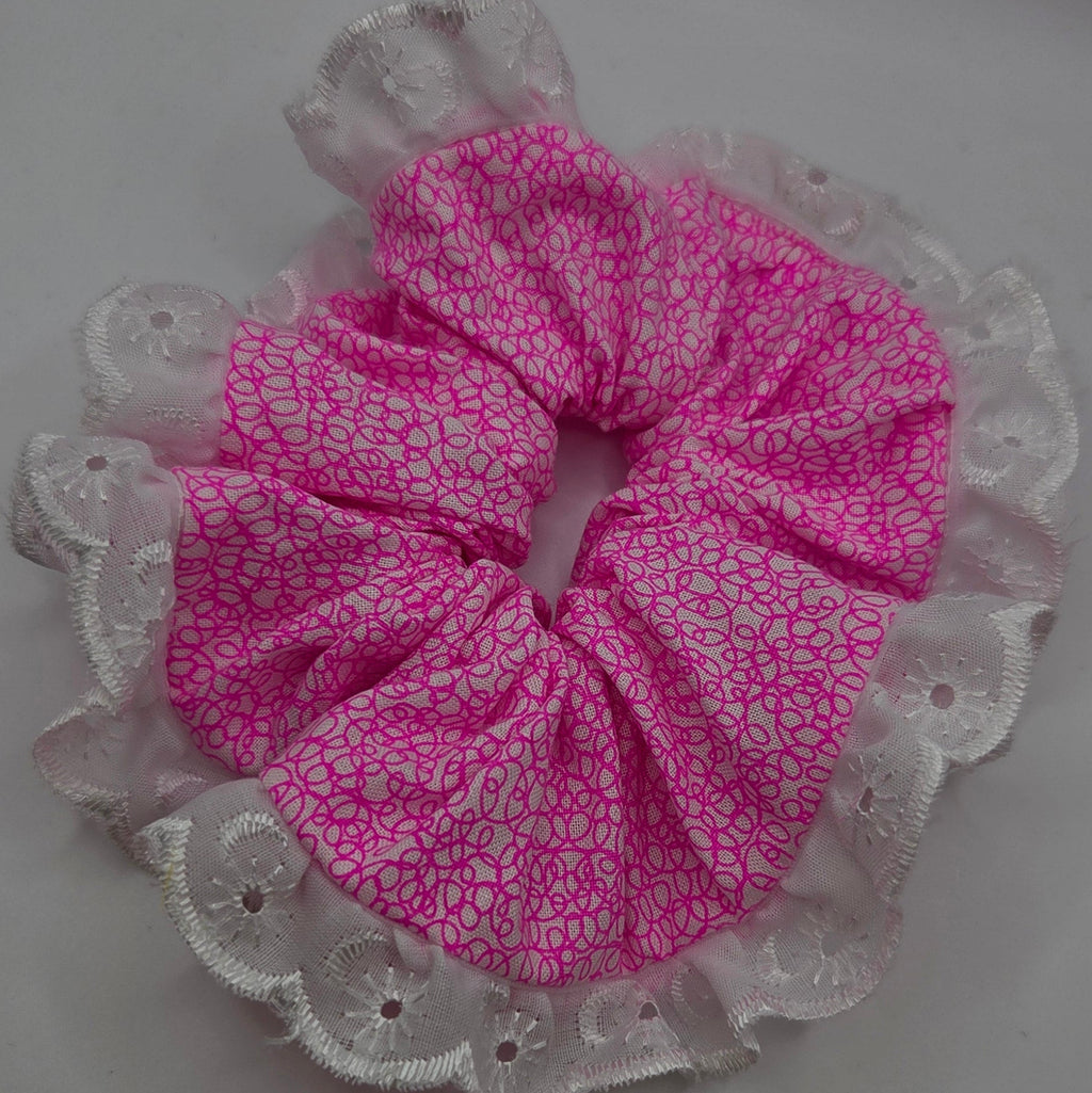 A pink fabric hair scrunchie with a white floral pattern and a white lace trim, shown on a plain light gray background.